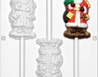May include: A clear plastic candy mold featuring three cavities for lollipops. Two cavities are empty, showing the embossed shapes of a Santa Claus and a gift sack. One cavity is filled with a colorful Santa Claus figure in red, white, and green.