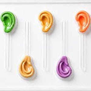 May include: A clear plastic mold tray featuring five distinct ear-shaped cavities. The mold contains five colorful ear-shaped objects in green, light orange, dark orange, tan, and purple, each attached to a clear plastic stick handle.