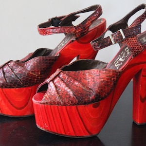 May include: Pair of vintage platform sandals with red wooden soles and heels. The uppers are made of a snakeskin-like material in shades of brown and red. Ankle straps with buckles complete the design. The heels measure approximately 4 inches.