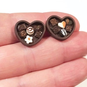 May include: Two miniature heart-shaped boxes filled with assorted chocolate candies. The boxes are brown and have a glossy finish. The candies are various shapes and colors, including brown, white, and orange.