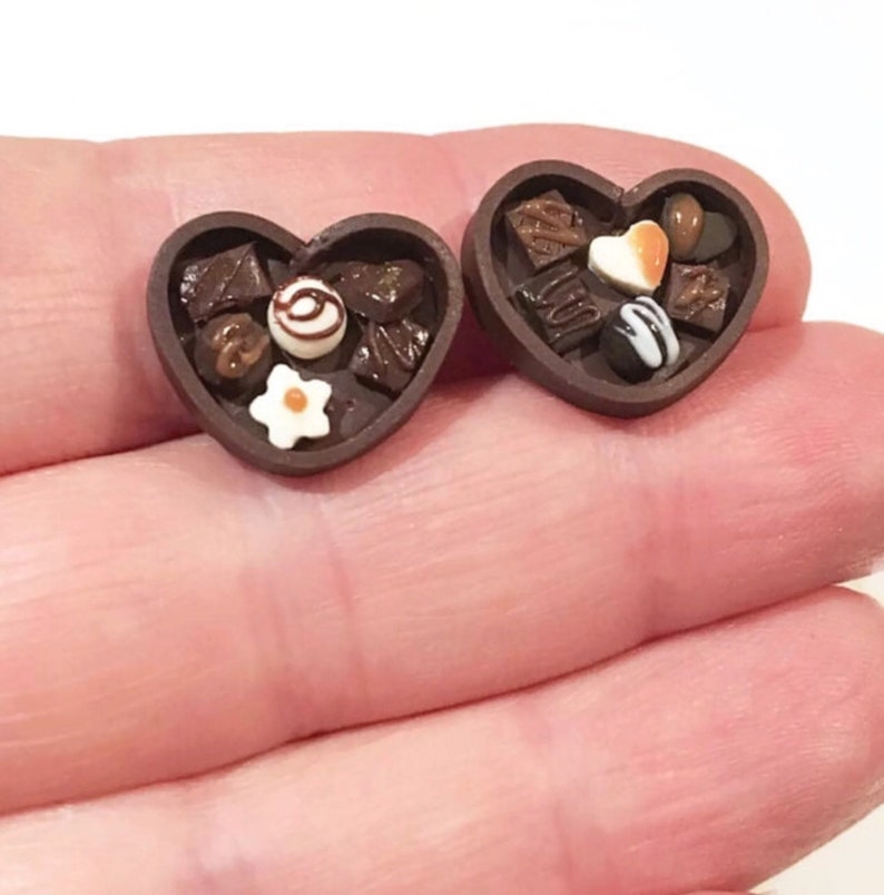 May include: Two miniature heart-shaped boxes filled with assorted chocolate candies. The boxes are brown and have a glossy finish. The candies are various shapes and colors, including brown, white, and orange.