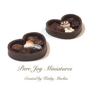 May include: Two miniature heart-shaped boxes filled with assorted chocolate candies. The boxes are made of dark brown chocolate and have a glossy finish. The candies are a variety of shapes and sizes, including squares, circles, and hearts. Some of the candies are decorated with white and orange accents.