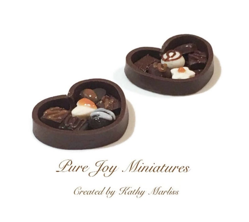 May include: Two miniature heart-shaped boxes filled with assorted chocolate candies. The boxes are made of dark brown chocolate and have a glossy finish. The candies are a variety of shapes and sizes, including squares, circles, and hearts. Some of the candies are decorated with white and orange accents.