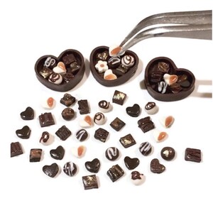 May include: A collection of miniature chocolate candies in various shapes, including hearts, squares, and swirls, are scattered around three heart-shaped boxes. The boxes are filled with more chocolates and are made of dark brown clay.