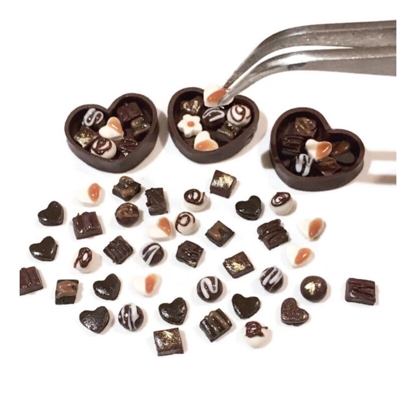 May include: A collection of miniature chocolate candies in various shapes, including hearts, squares, and swirls, are scattered around three heart-shaped boxes. The boxes are filled with more chocolates and are made of dark brown clay.