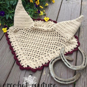 May include: A crocheted horse fly bonnet in beige with a burgundy trim. The bonnet is laying on a wooden surface with two horseshoes.