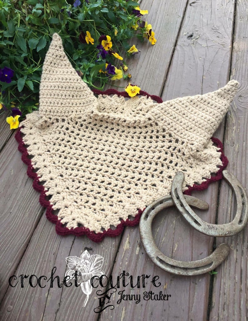 May include: A crocheted horse fly bonnet in beige with a burgundy trim. The bonnet is laying on a wooden surface with two horseshoes.