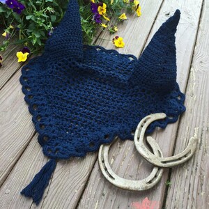 May include: A navy blue crocheted horse fly bonnet with a tassel. The bonnet is laying on a wooden surface with two horseshoes.