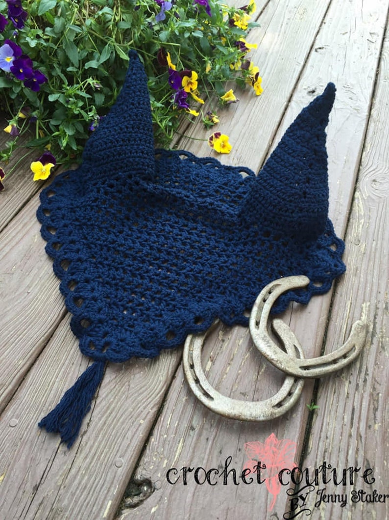 May include: A navy blue crocheted horse fly bonnet with a tassel. The bonnet is laying on a wooden surface with two horseshoes.