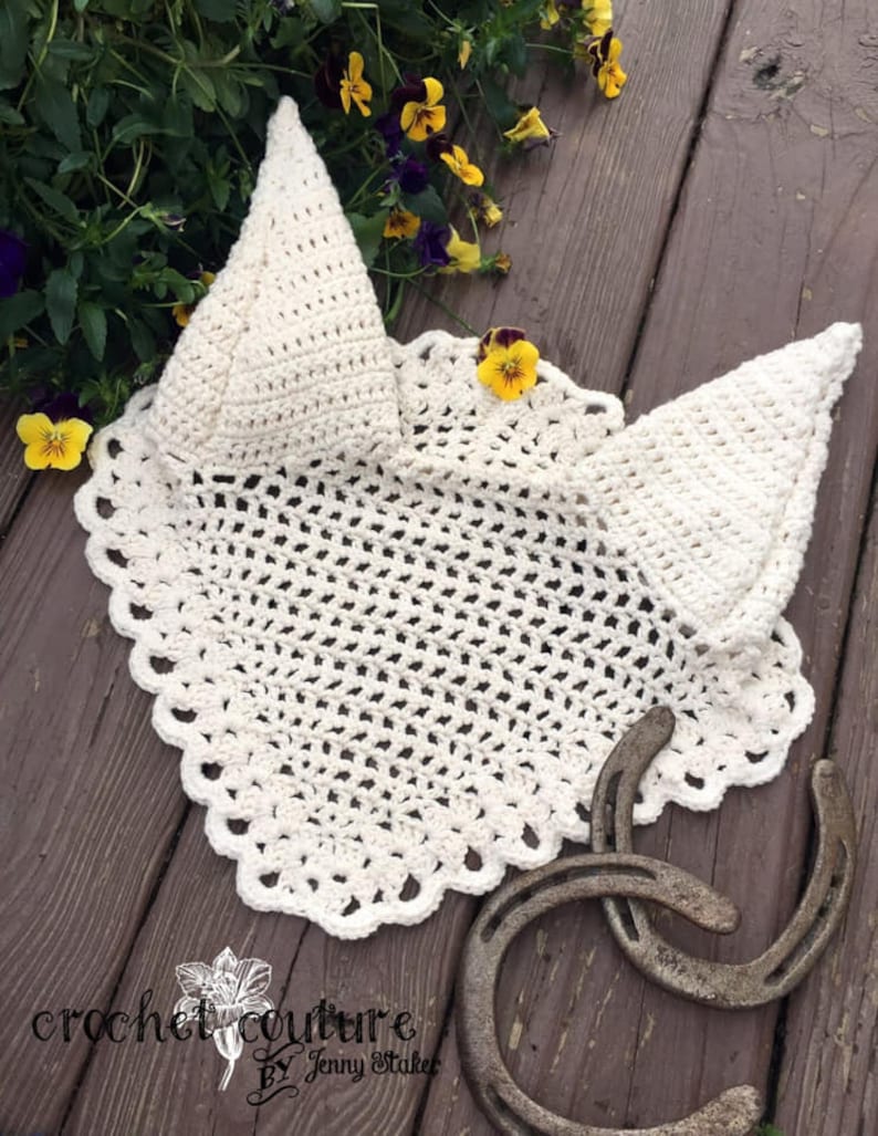 May include: A crocheted horse fly bonnet in a cream color with a scalloped edge. The bonnet is laying on a wooden surface with two horseshoes.