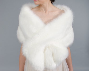 May include: White faux fur shawl with a soft, fluffy texture. The shawl is draped over the shoulders of a person wearing a white dress.