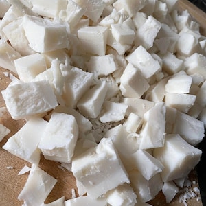 May include: A close-up of a wooden cutting board covered in small, white, cubed pieces of coconut.