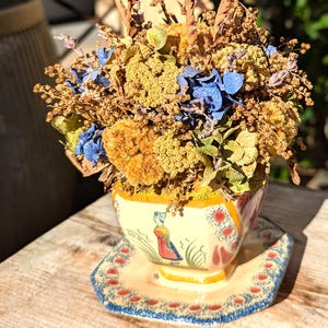 May include: A dried flower arrangement in a decorative ceramic cup and saucer. The cup is yellow with a painted figure and floral designs. The arrangement includes blue, yellow, and brown dried flowers. The saucer has a blue and red border.