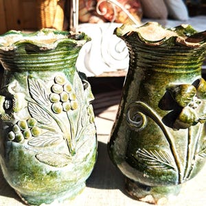 May include: Two green ceramic vases with textured surfaces and ruffled rims. One vase features a floral design with leaves and berries, while the other showcases a large flower and swirling accents. The vases are likely for decorative purposes.