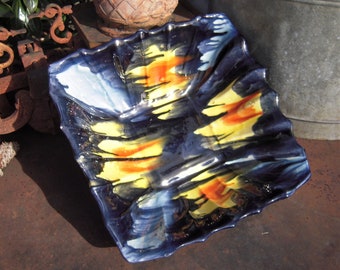 May include: A square, ceramic serving dish with a wavy edge. The dish is primarily navy blue with abstract brushstrokes of yellow, orange, and white. The dish is likely used for serving food or as a decorative piece.