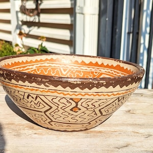 May include: A round ceramic bowl featuring a hand-painted geometric pattern in shades of dark brown, terracotta orange, and cream. The exterior and interior rim are decorated with repeating zigzag lines and intricate angular maze-like motifs.