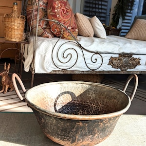 May include: A large, antique copper bowl with two curved handles. The bowl has a hammered texture and a dark, aged patina. The background includes a wrought iron daybed with patterned pillows and a wicker-covered bottle.
