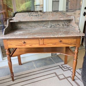 May include: Antique wooden desk with a marble top. The desk features two drawers with round knobs, a lower shelf, and ornate, turned legs. The marble top has a reddish-brown and gray pattern. The desk is a classic piece of furniture.
