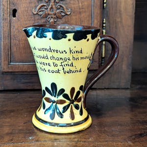 May include: A cream-colored ceramic pitcher with a dark brown handle and rim. The exterior features hand-painted dark brown and black floral motifs at the base and black cursive text that reads: is us wondrous kind, that would change his mind, he were to find, in his coat behind.