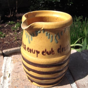 May include: A yellow ceramic pitcher with a spout and a wide opening. The pitcher has dark brown horizontal stripes and the words "le coup club doux" in brown. It has green drips near the top.