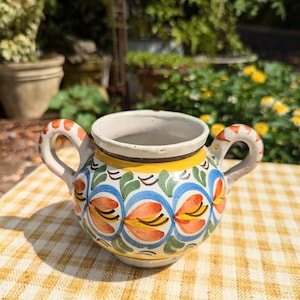 May include: A small, hand-painted ceramic jar with two handles. The jar features a white interior, a yellow band, and a floral design in orange, blue, and green. The handles have an orange and white pattern. It sits on a yellow checkered tablecloth.