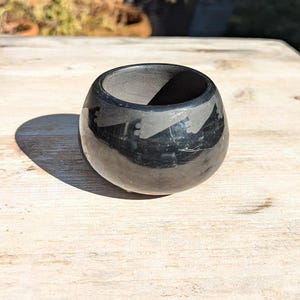 May include: A small, round, black ceramic bowl with a glossy finish. The bowl has a wide opening and a smooth interior. The exterior reflects the surrounding environment. The bowl is sitting on a light-colored wooden surface.