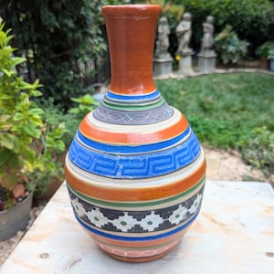 May include: A hand-painted ceramic vase with a bulbous body and a narrow neck. The vase features a terracotta-colored neck and a body adorned with horizontal bands of blue, orange, green, and black geometric patterns. The vase is approximately 10 inches tall.