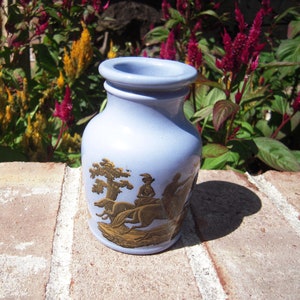 May include: A light blue ceramic jar with a gold design of people riding horses. The jar has a rounded body and a wide mouth.