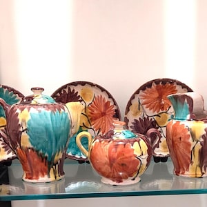 May include: A collection of hand-painted ceramic tableware featuring a floral design. The set includes teacups, saucers, plates, teapots, and a creamer. The color palette includes teal, orange, brown, and yellow. The pieces are displayed on a glass shelf.