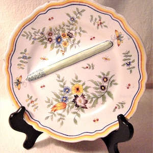 May include: A white ceramic plate with a scalloped edge and a green asparagus design. The plate is decorated with yellow, red, and blue flowers and green leaves. The plate is on a black stand.