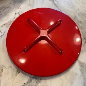 May include: A circular, glossy red lid featuring a raised, cross-shaped handle centered on its top surface. The lid has a smooth, reflective finish and a simple, clean design, providing a solid cover for a corresponding container or pot.