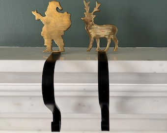 May include: Two Christmas stocking holders. One is a gold-colored Santa Claus silhouette, the other a gold-colored reindeer. Both have black metal hooks for hanging stockings. The holders are displayed on a white marble surface.