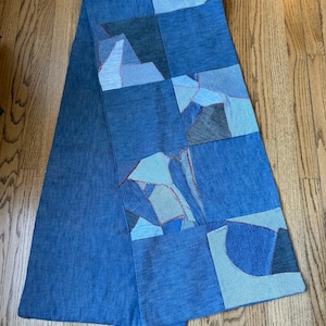 May include: A blue denim patchwork scarf with different shades of blue and gray denim pieces sewn together. The scarf is folded in half and laid on a wooden floor.