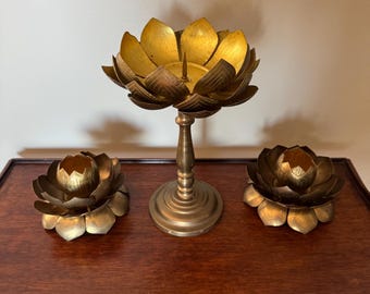 May include: Three brass lotus flower candle holders. The largest is a tall, stemmed candle holder. Two smaller lotus flower candle holders are on either side. The candle holders are gold in color and sit on a dark wooden surface.