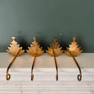 May include: A set of four gold-toned metal stocking holders, each featuring a flat, stylized Christmas tree silhouette topped with a small star. Each tree is attached to a long, slender metal hook with a curved end for hanging stockings.