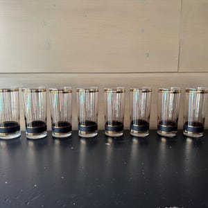 May include: Eight clear glass tumblers with black and gold trim. The glasses are lined up in a row on a black surface.