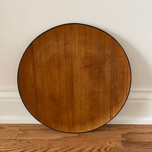 May include: A round wooden tray with a dark rim. The tray has a natural wood grain pattern in shades of brown and tan. The tray is set against a white wall and a wooden floor.