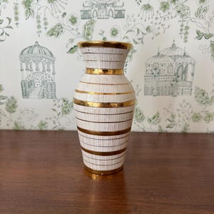 May include: A white ceramic vase featuring a series of thin, vertical brown lines and horizontal gold bands. The vase has a flared rim with a gold-painted edge and a tapered neck that widens toward the base, creating a balanced, symmetrical silhouette.