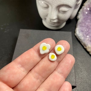 Puede incluir: Tres pendientes de cerámica blanca con forma de huevos fritos con yemas amarillas. Los pendientes se muestran en una mano.
