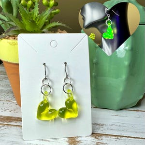 Pode incluir: Um par de brincos pendentes com ganchos prateados e pingentes em forma de coração verde. Os pingentes são translúcidos e têm um leve tom amarelo.