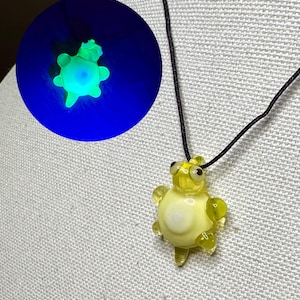May include: A yellow glass turtle pendant with a black cord. The turtle has a white belly and clear flippers. A separate image shows the turtle glowing green under a black light.