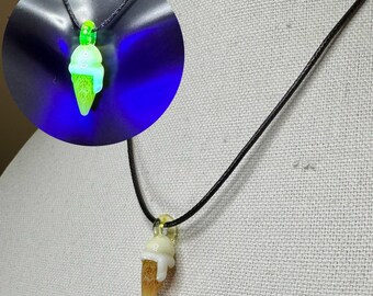 May include: A black cord necklace with a glass ice cream cone pendant. The pendant is white with a yellow and brown cone and a green top. The pendant glows green under a black light.
