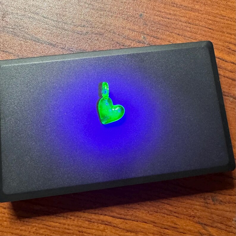 May include: A small, green glass heart pendant with a silver bail. The pendant is glowing under a blacklight.