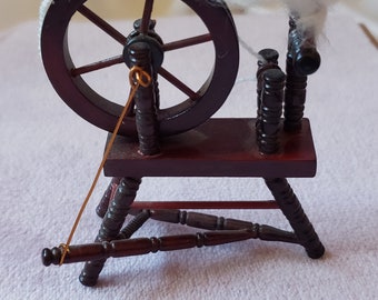 May include: Miniature wooden spinning wheel with a dark brown finish. The wheel has a large circular wheel and a small platform. A mass of fluffy white fiber sits atop the wheel. A copper-colored wire is attached to the wheel.