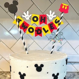 May include: A white birthday cake with red frosting and black Mickey Mouse heads. The cake is decorated with black and gold sprinkles. A cake topper with a banner that reads 'Oh Twodles' is on top of the cake.