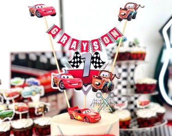 May include: A two-tiered birthday cake decorated with a red, white, and black checkered pattern. The top tier features a red and white racing car design with the number "1" on it. The cake is topped with a red race car figurine and a banner that reads "GRAYSON".