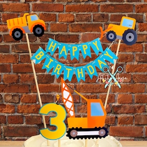 May include: A white cake with blue frosting decorated with a construction-themed cake topper. The topper includes a banner that says "Happy Birthday", a number 3, and three construction vehicles.