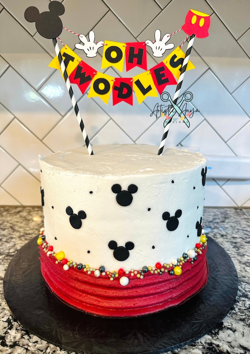 May include: A white birthday cake with red frosting and black Mickey Mouse heads. The cake is decorated with black and gold sprinkles. A cake topper with a banner that reads 'Oh Twodles' is on top of the cake.