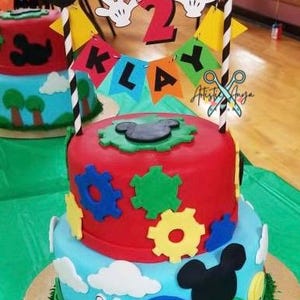 Pu&ograve; includere: Torta di compleanno a due piani decorata con personaggi di Topolino, ingranaggi e uno striscione che recita "Klay 2". La torta &egrave; glassata in rosso, blu e verde.