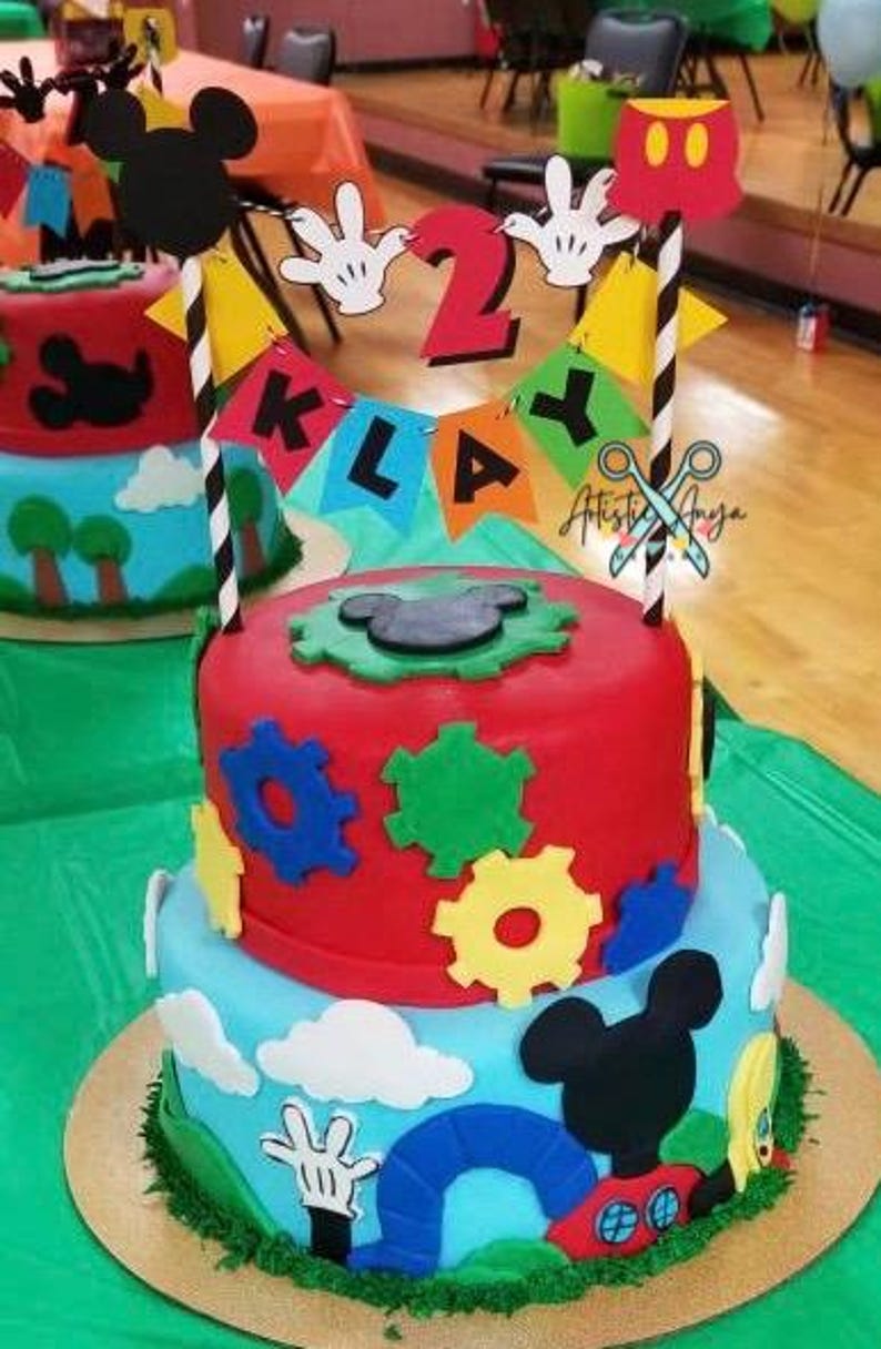 Pu&ograve; includere: Torta di compleanno a due piani decorata con personaggi di Topolino, ingranaggi e uno striscione che recita "Klay 2". La torta &egrave; glassata in rosso, blu e verde.